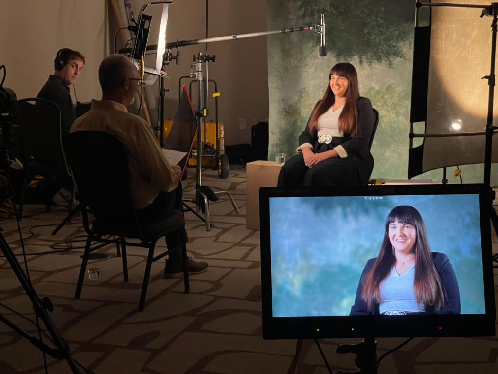 A behind-the-scenes view of a video interview setup, featuring a woman with long dark hair seated in front of a textured green background, lit by professional lighting. A monitor displays her on-screen image, while a man with a script and another individual with headphones are seen managing the recording equipment.