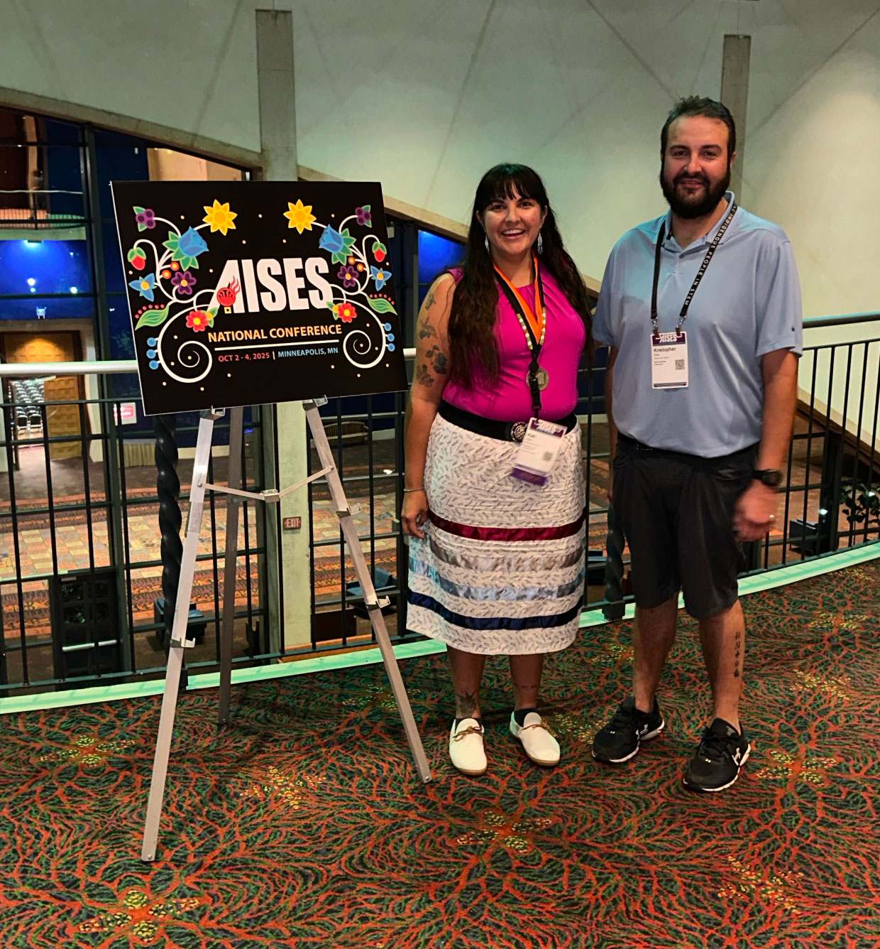 Two people standing indoors next to an easel displaying a colorful sign for the AISES National Conference, which features floral designs. The woman is wearing a bright pink top and a traditional ribbon skirt, while the man is dressed in a light blue shirt and shorts. They are smiling in a well-lit conference venue.