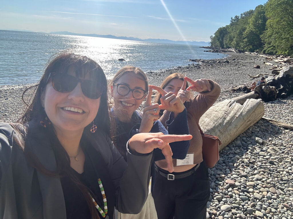 Three women smiling and posing for a selfie on a sunny pebble beach, with one wearing sunglasses and beaded jewelry. Behind them, a driftwood log lies on the shore, and the shimmering water and distant mountains are visible under a clear blue sky.